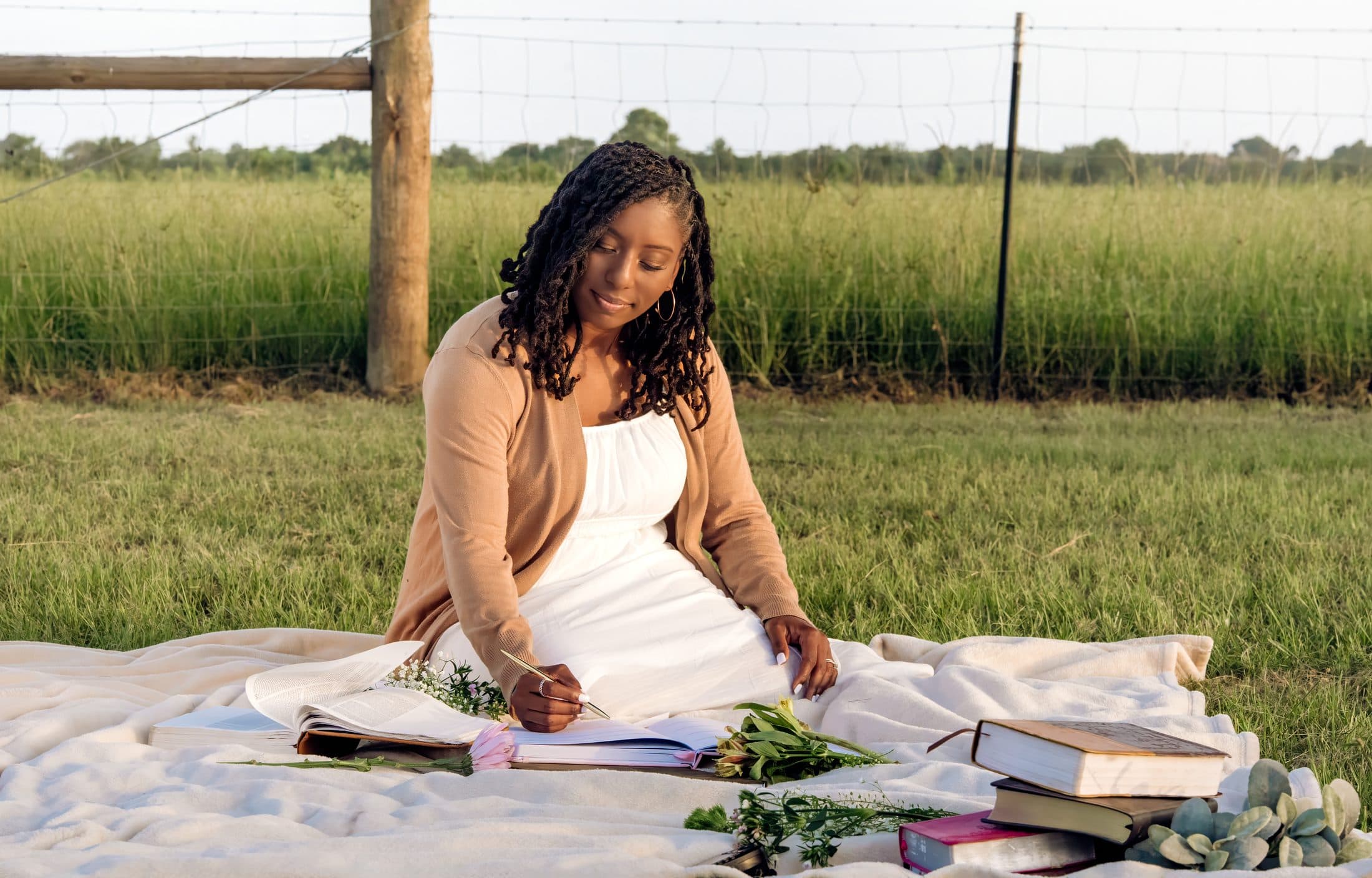 A woman journaling in the Ezer Planner surrounded by flowers and books on a blanket