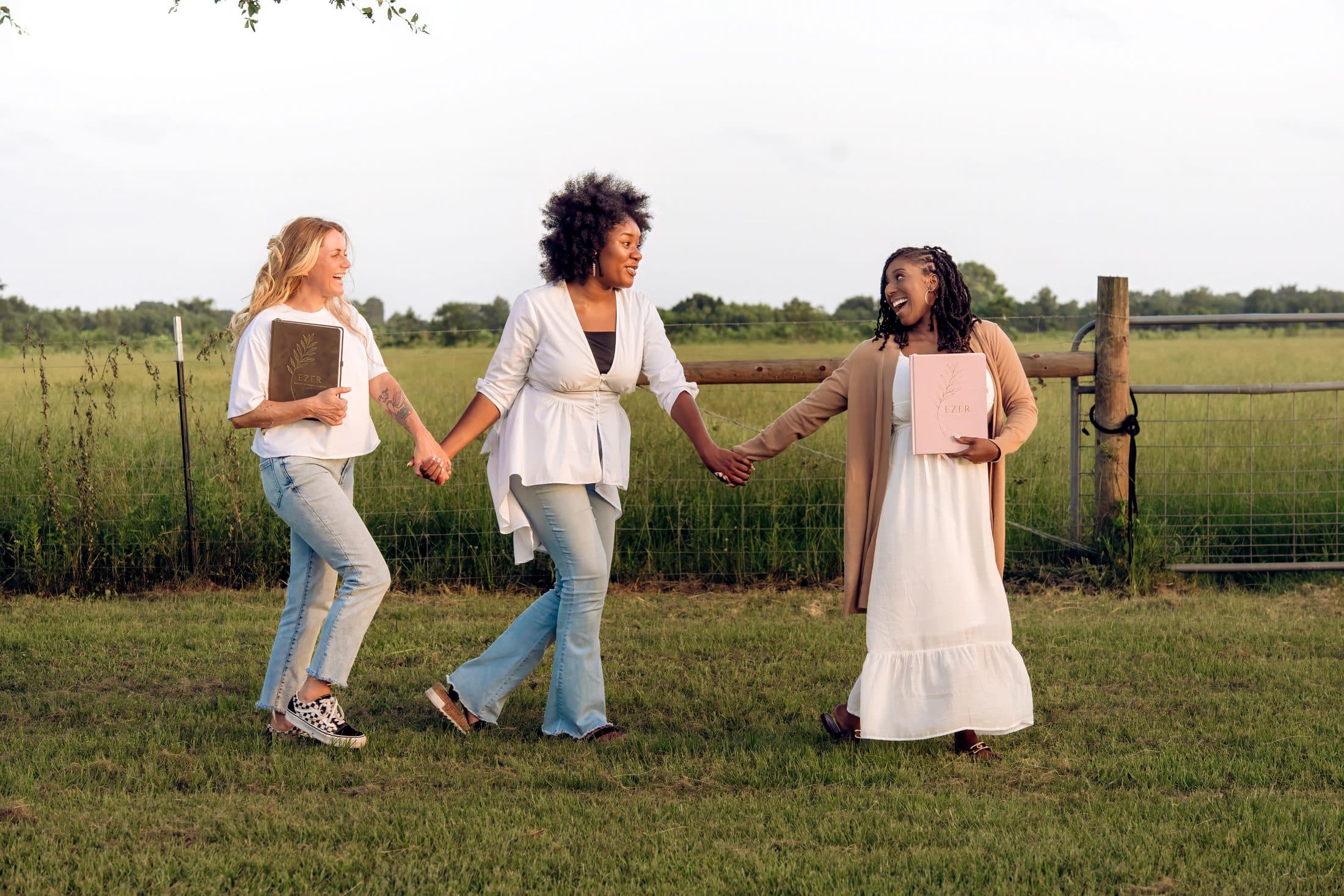 Three women walking hand in hand through a field, each holding an Ezer Planner